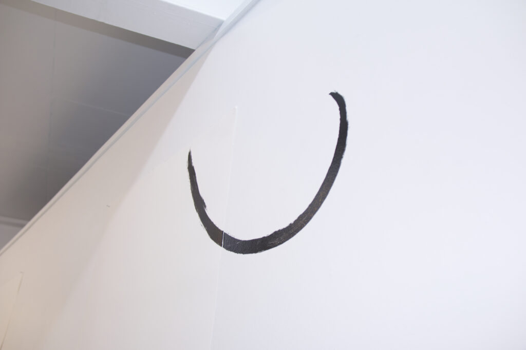 A printed black line curved like a half moon, printed on a gallery wall and across a sheet of hung paper. 