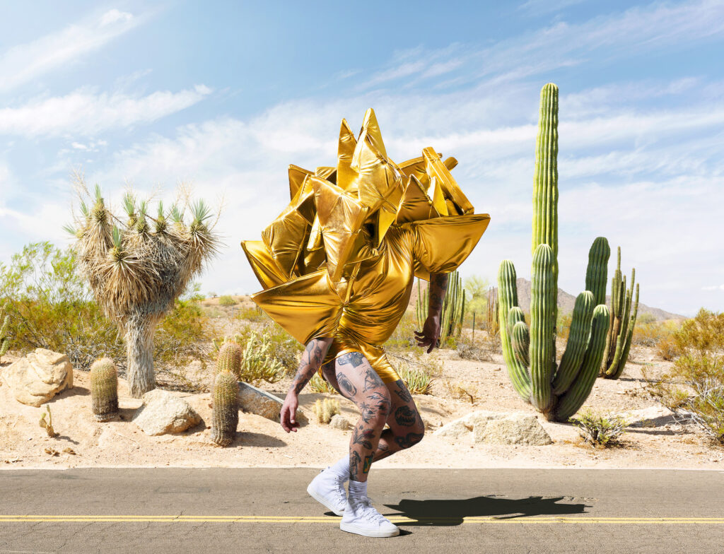 A person in an angular gold costume, awkwardly poised as if frozen in a slow race. Cacti and desert plants behind.