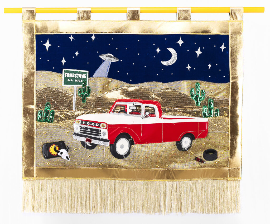 A textile banner featuring a red and white Ford pick up truck, in the desert on the way to Tombstone, alien spacecraft beaming from the night sky above, surrounded by cacti, skulls, and car parts. 