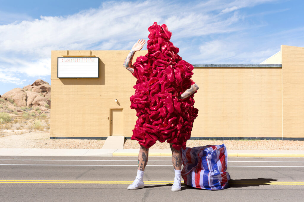 A person in a red costume, waving in front of a dilapidated theatre, a red, white and blue storage bag by their side.