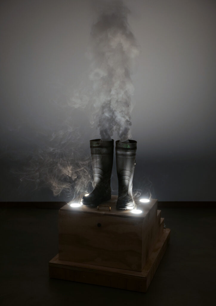 Two black rubber boots sit atop a wooden platform, smoke floats out from the boots and fills the room with a haze.