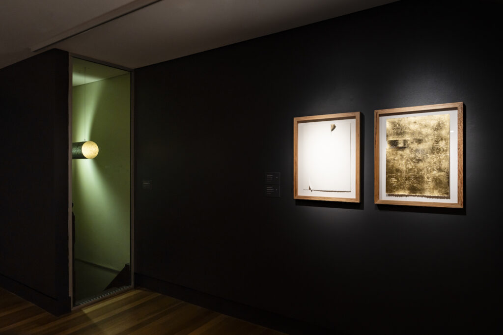A darkened gallery space has a glowing object in a doorway and two works, white and gold, in timber frames.