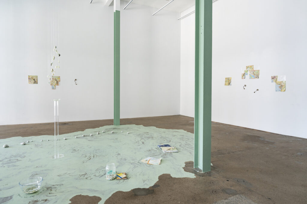 Two sea-green pillars stretch to the ceiling of a white-walled gallery room. Green paint spills out between the pillars, across the concrete floor. There are small street maps and wires wrapped with the artist’s hair pinned to the walls, a chandelier of pearls and stones hanging in the centre of the room above a clear resin pillar, and a line of small paper stones, map pages, and vessels of water placed on the floor.