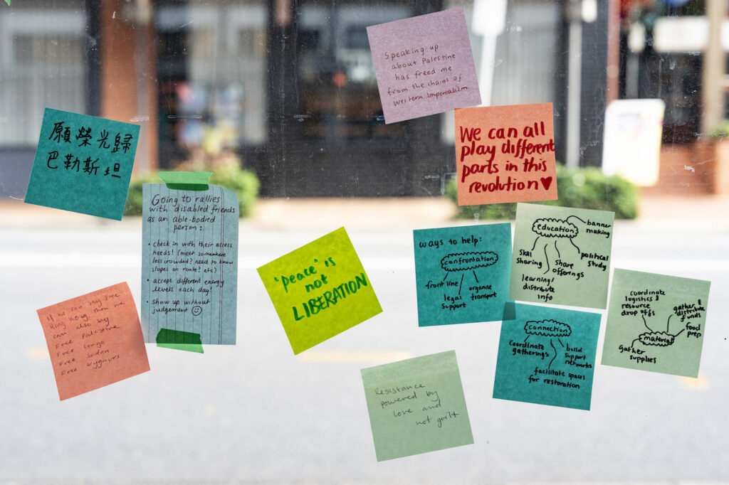 Multi-coloured post-it notes are fixed to a gallery window, with a view of the street beyond. The post-it notes contain handwritten messages sharing knowledge, hope, and support for Palestine.