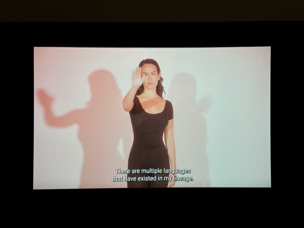 Projected video still of the artist with their right hand raised outward to the screen. Subtitled text at the bottom of the screen reads: “There are multiple languages that have existed in my lineage.”