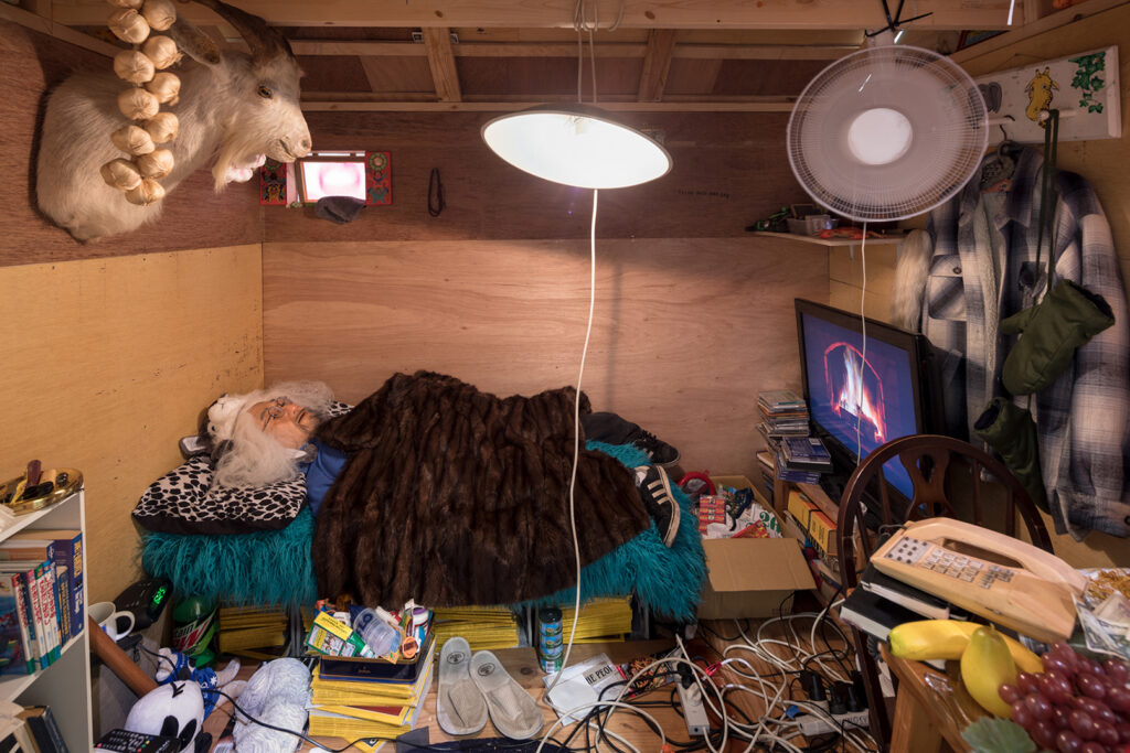 Interior view of a tiny house. Against the wall, a model of the artist as an elderly man sleeps beneath a blanket, surrounded by magazines, toys, electrical cords, medicines, and other everyday objects.