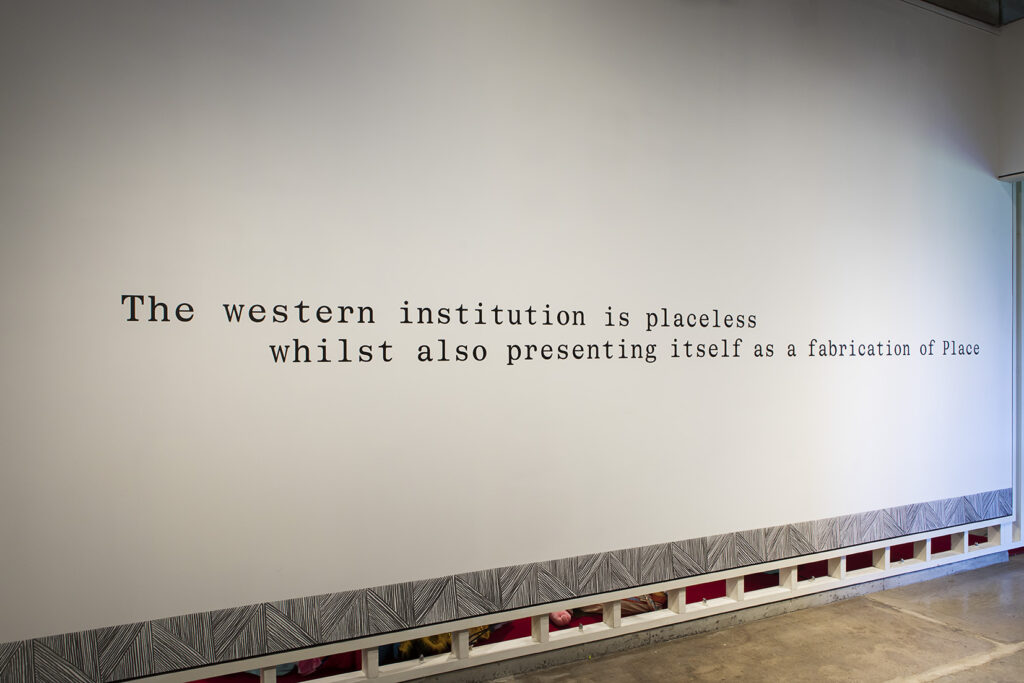 Text on a gallery wall reads: “The western institution is placeless | whilst also presenting itself as a fabrication of Place.” A repetitive line drawing features along the bottom of the wall.