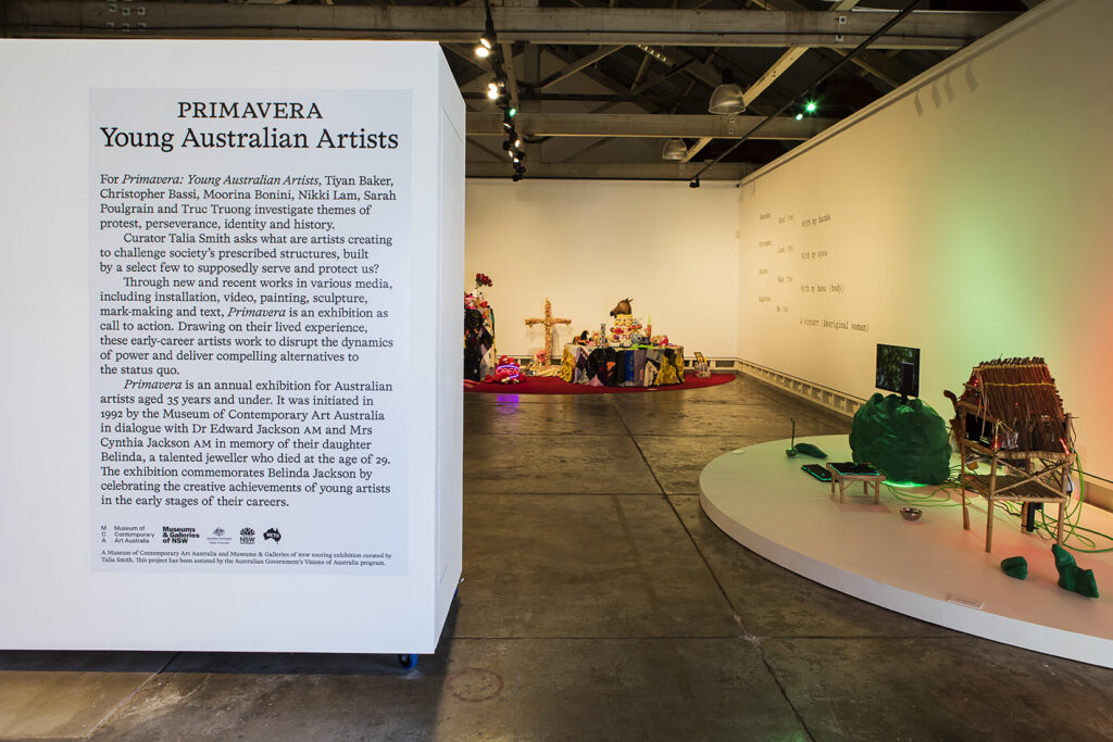 A photograph of a white gallery with introductory text on a nib wall to the left and three contemporary artworks to the right.