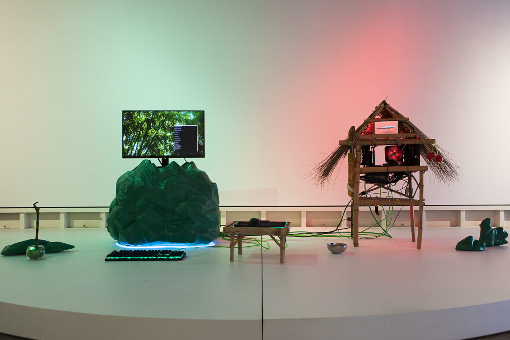 An installation artwork in a gallery featuring a screen, a small hand-made house, and other diverse elements.