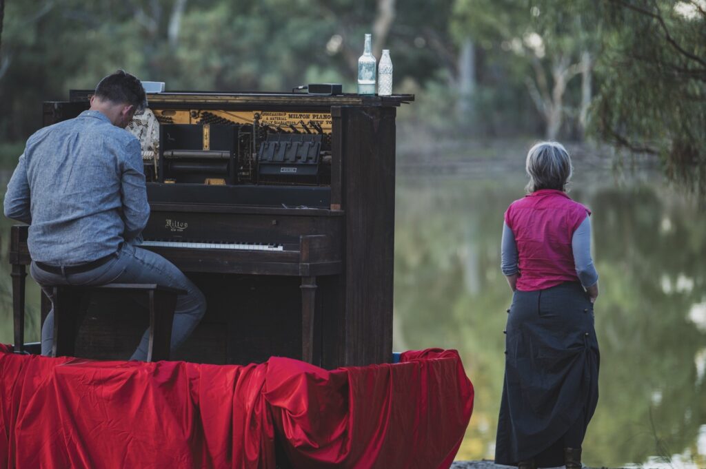 A man on a raised red stage plays a piano, a woman standing nearby looks into the Australian landscape.