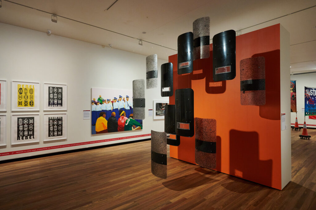 In a gallery, riot shields hang in front of an orange painted wall; further works hang in the background.