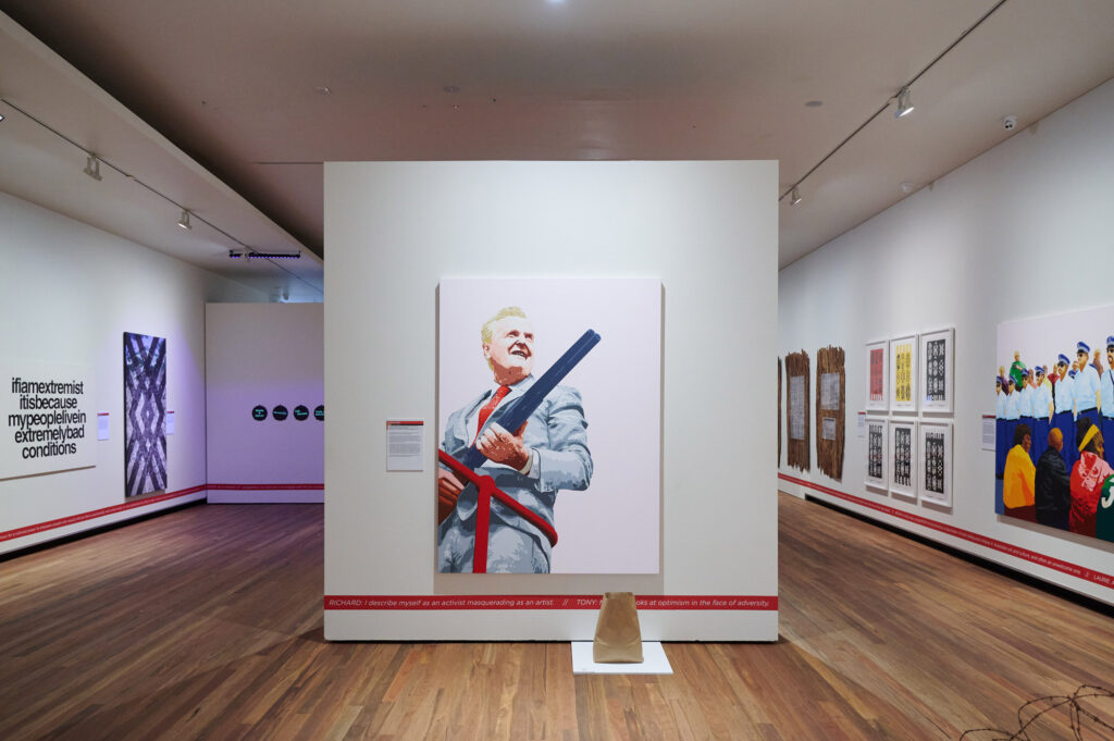 In a gallery, a portrait of a white man wearing a suit and holding a gun hangs on a partition wall; further works hang on the main walls either side.