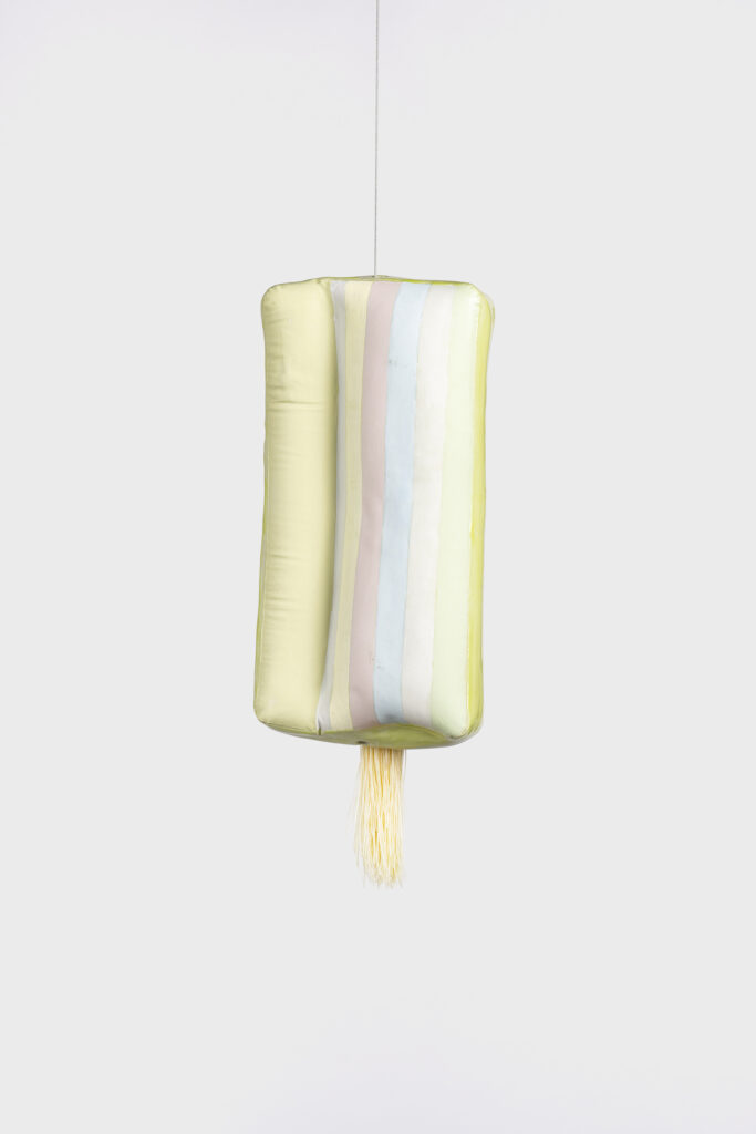 A photograph of a suspended ceramic form, glazed in a spectrum of greens and blues, with a light yellow tassel adornment. 