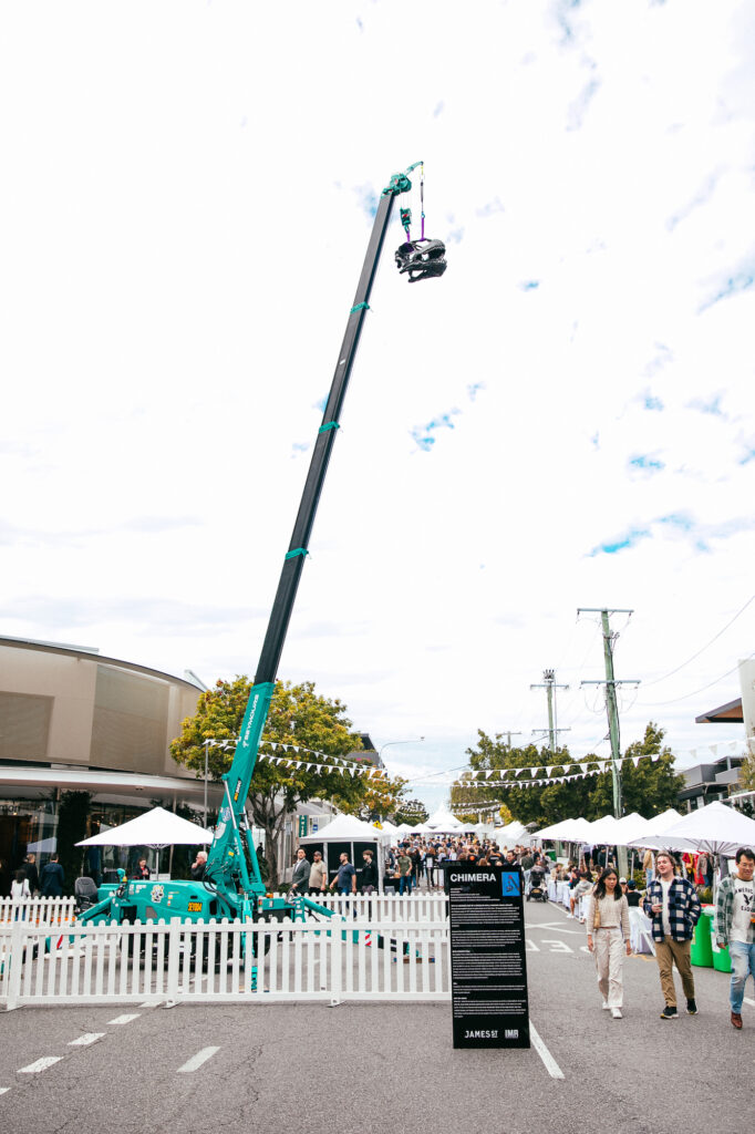 Amidst a street festival, a dinosaur skull replica hangs over the crowd, lifted by a crane.