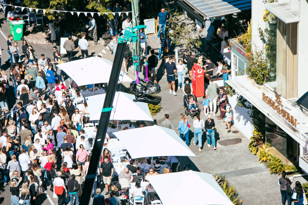 Amidst a street festival, a dinosaur skull replica hangs over the crowd, lifted by a crane.