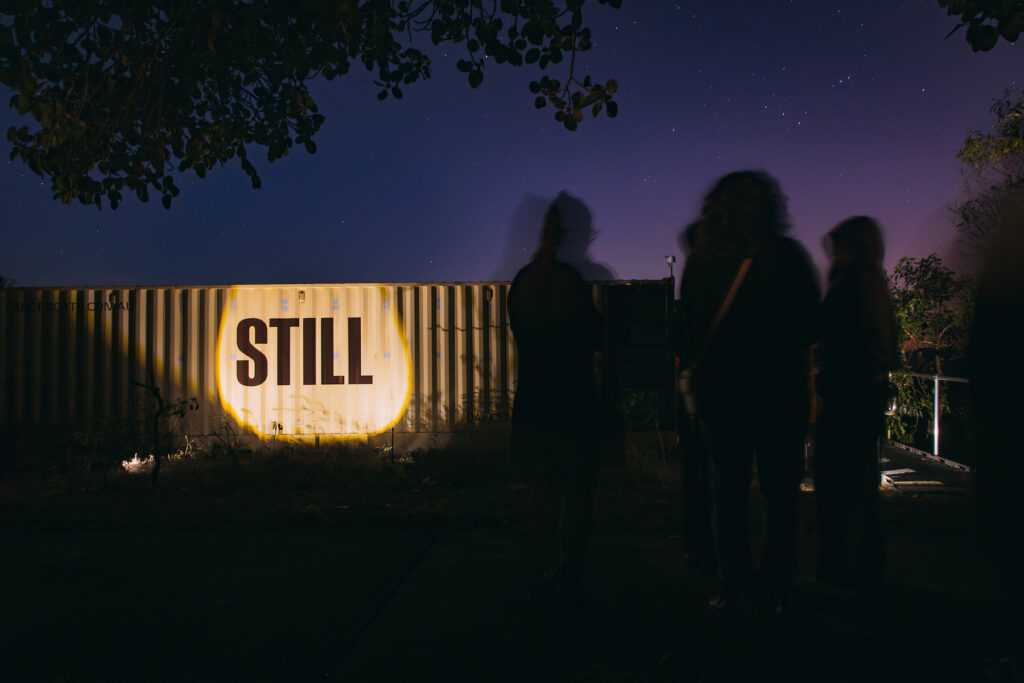 A photograph of figures moving in the dark, behind them is a shipping container with the large word STILL lit by a spotlight.