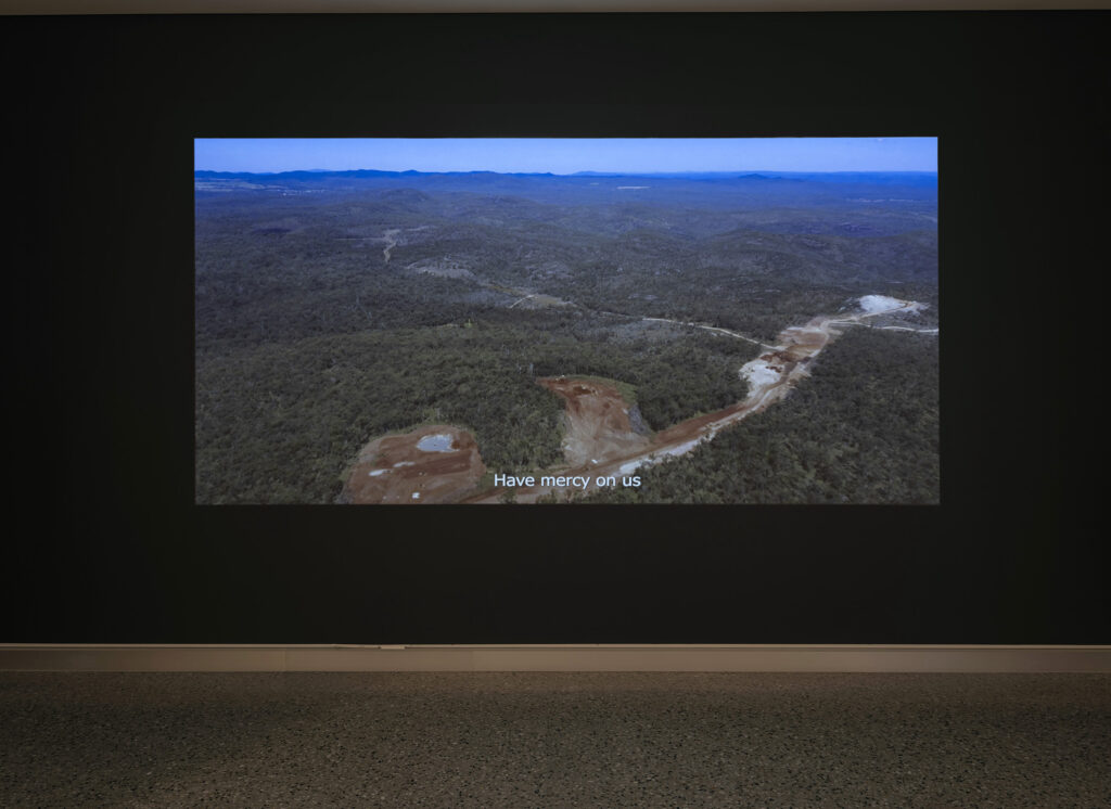 A film plays in a darkened gallery; text reads “Have mercy on us” above an Australian landscape with a road in development.