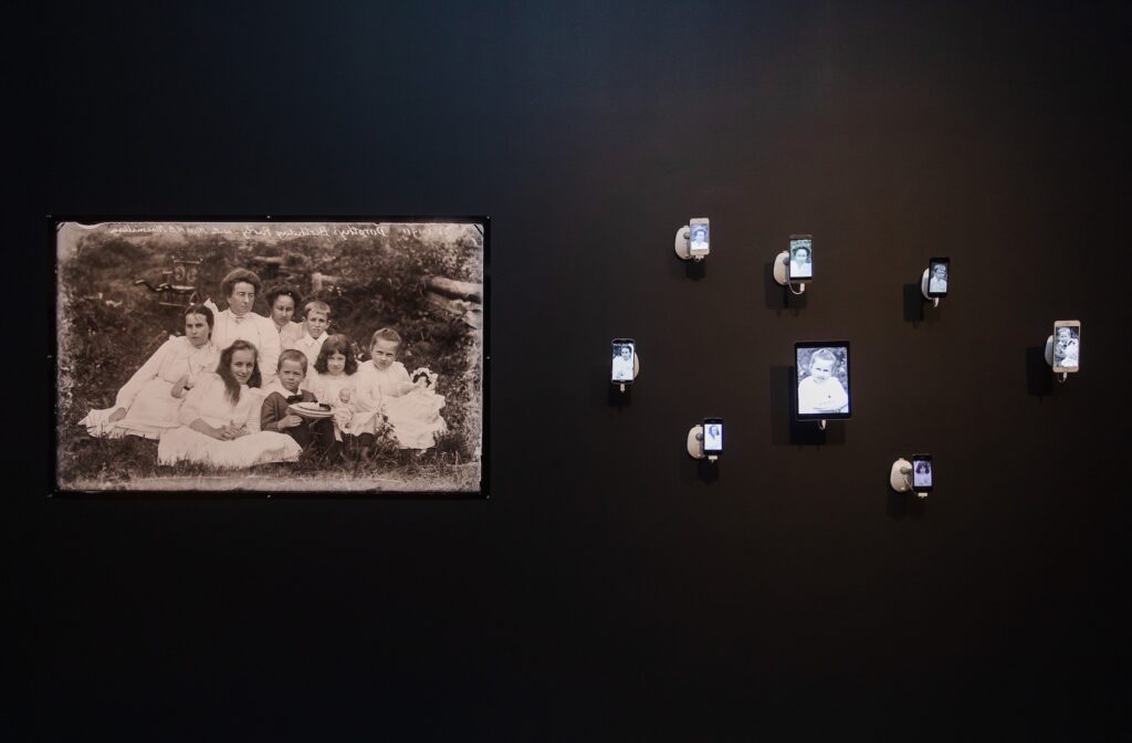 A large archival photograph of a family hangs on a gallery wall. To the right, their faces are excised and rearranged on smart phones.