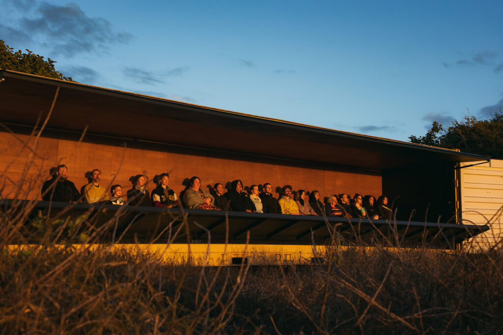 An audience sitting in a constructed box in nature bask in the rising sun’s glow.