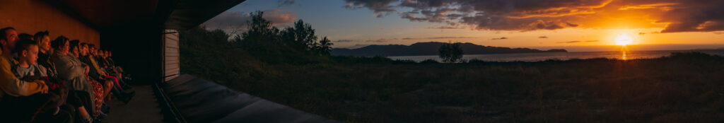 A panoramic photograph with audiences sitting in a shipping container to the left and the sunrise in nature to the right.