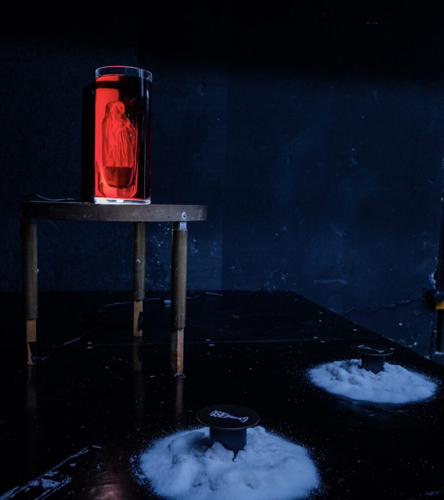 A sculpture of the Virgin Mary sits in red liquid on a raised platform, on the ground are two circular piles of salt, coasters propped up in the middle. The whole scene is set in a grimy dark club.