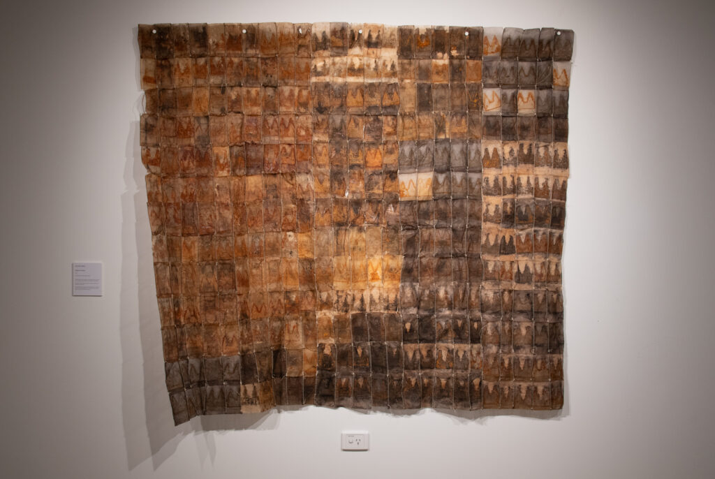 A patchwork quilt-like object of tea-stained browns hangs against a white wall.