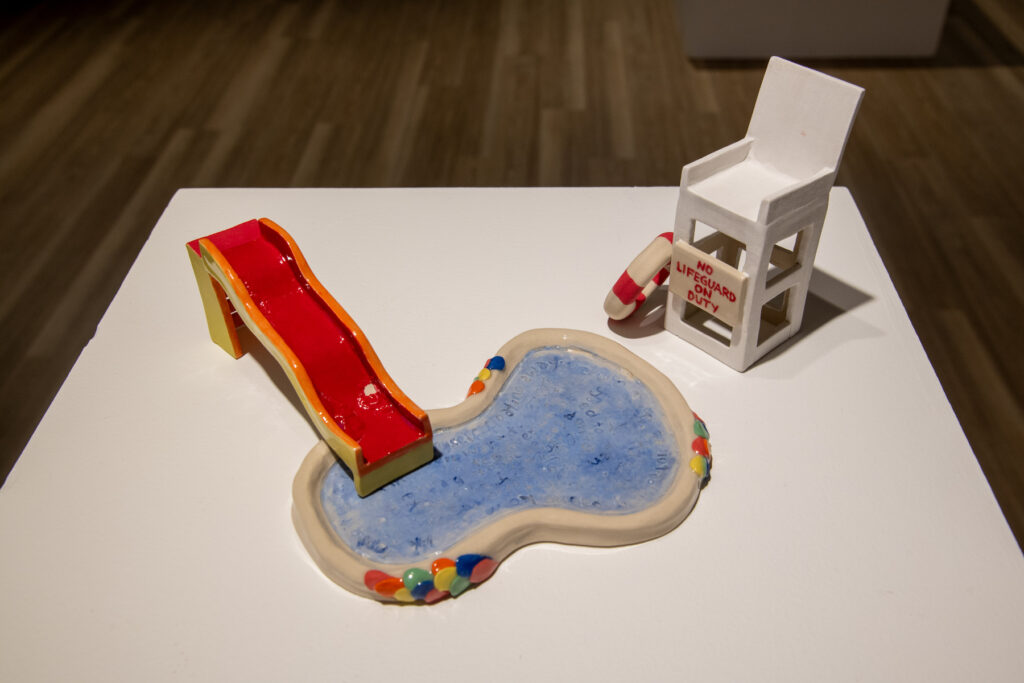 ID: A small ceramic pool with slide and lifeguard’s chair sit on a white plinth.