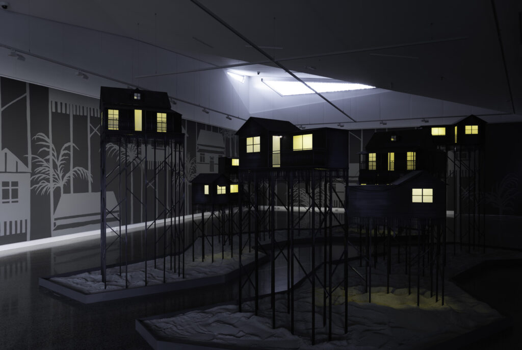 Dark sculptures of houses on stilts, glowing from within, fill a gallery.