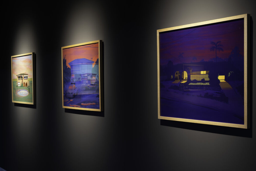 Three neon-coloured images of houses hang on a dark gallery wall.