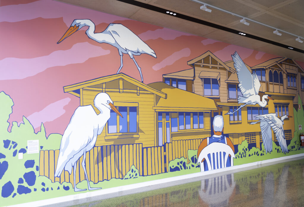 A mural depicts a figure sitting in a white plastic chair, gazing at a suburban house. Multiple large white birds fill out the scene.