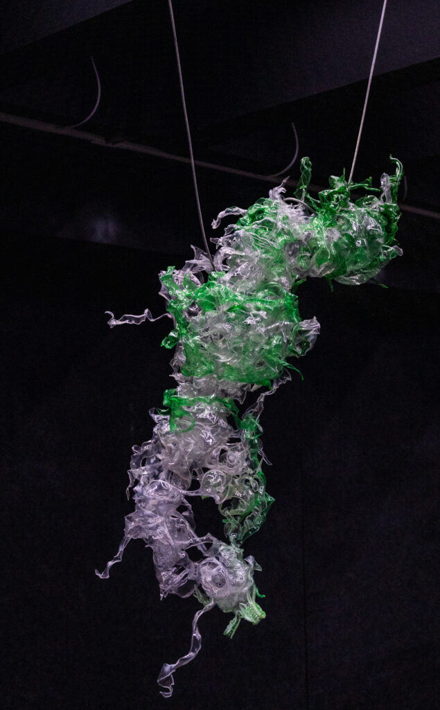 Twisted clear and green plastic forms hang from the ceiling in a dark space.