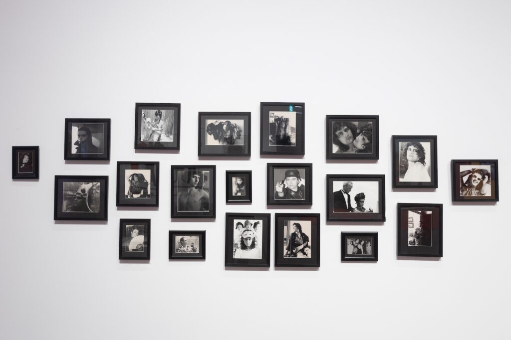 Black and white photographs of a young male figure in costumes fills a white gallery wall.