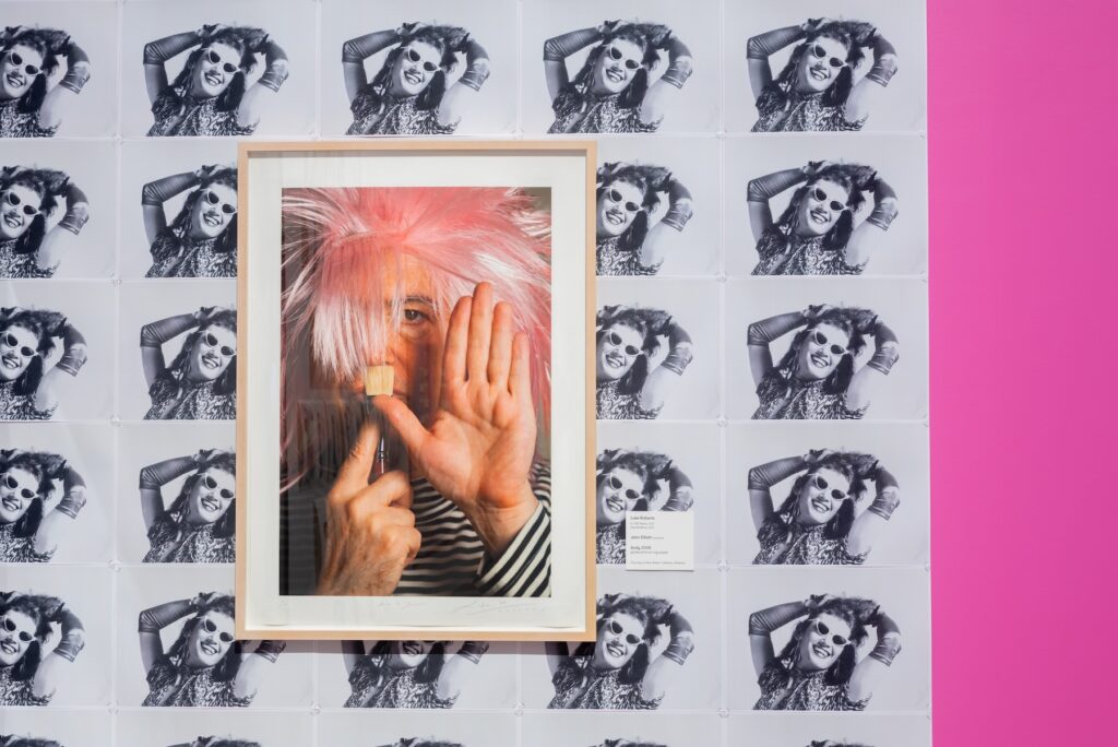 A photograph of a man wearing a light pink wig is flanked by repeated imagery of a black and white posed photograph and a bright pink wall.