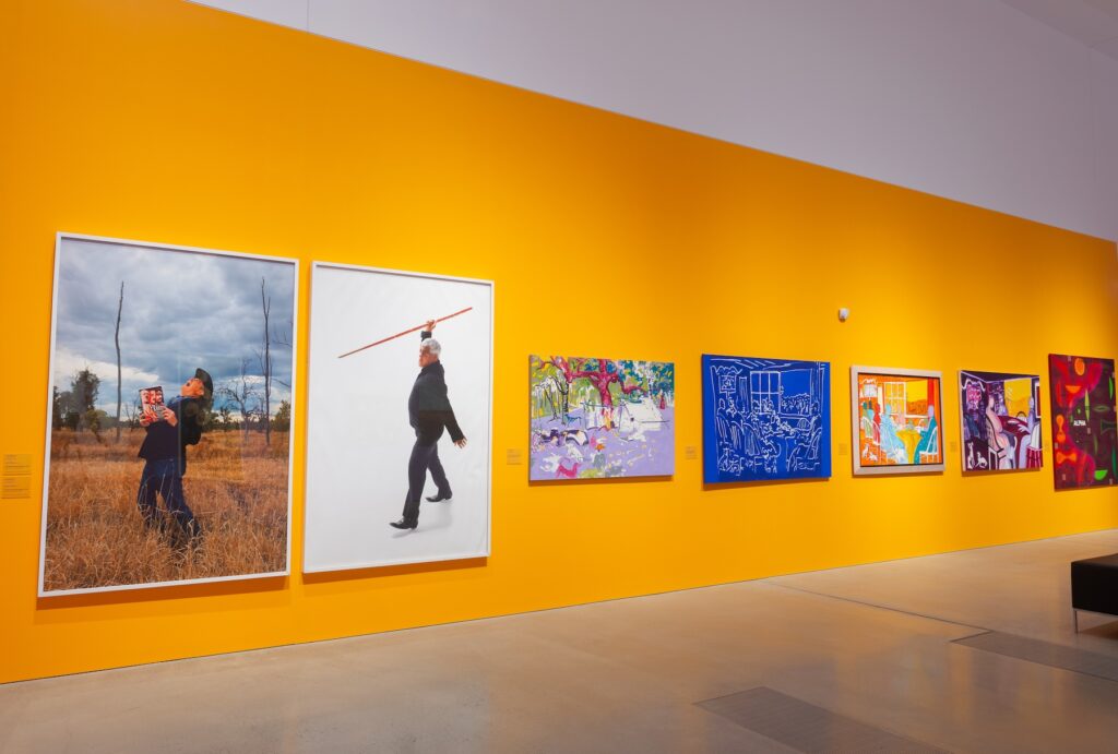  Large photographs and colourful paintings hang on an orange gallery wall.