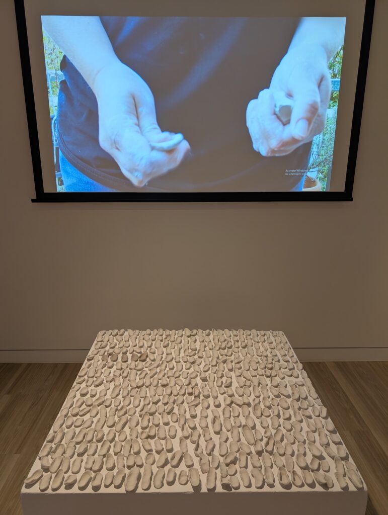 A video shows a close shot of a figure squeezing clay in their hands. In front of the video, 42 small clay shapes sit on a low plinth.