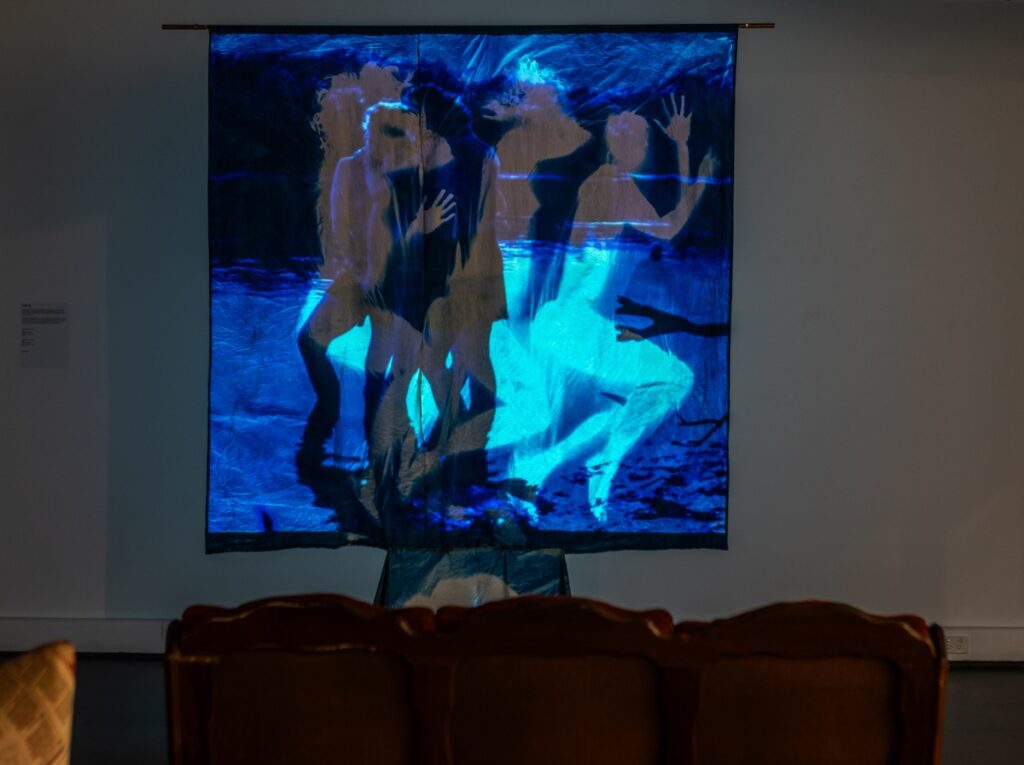 A photograph shows the back of a couch facing a projection screen. blue and white silhouettes of human figures overlap one another. There is the suggestion of a lake.