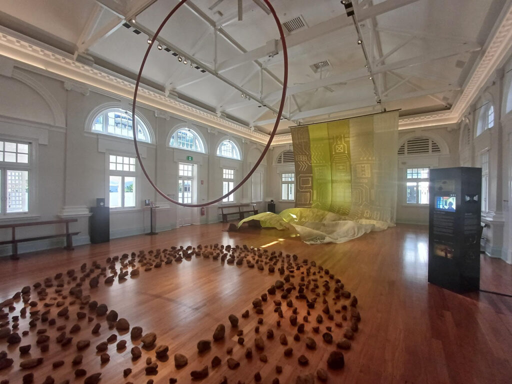 A colonial building transformed into a gallery shows rocks placed neatly in a circle on the ground, a hanging hoop and a large patterned textile.