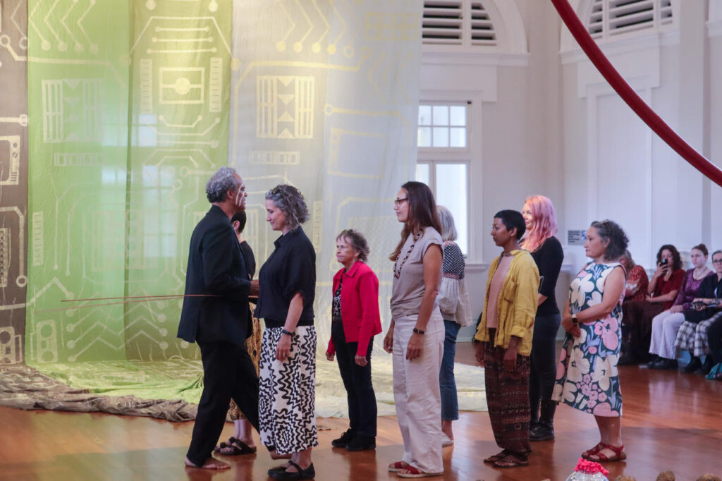 An Indigenous man stands in a gallery, a large length of textile behind him. In front of him is a group of Indigenous women, all facing him.