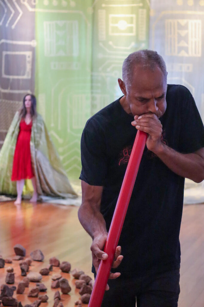 An Indigenous man plays a red yidaki (didgeridoo). Behind him are rocks placed on the ground, a woman in green and red, and a large textile work.