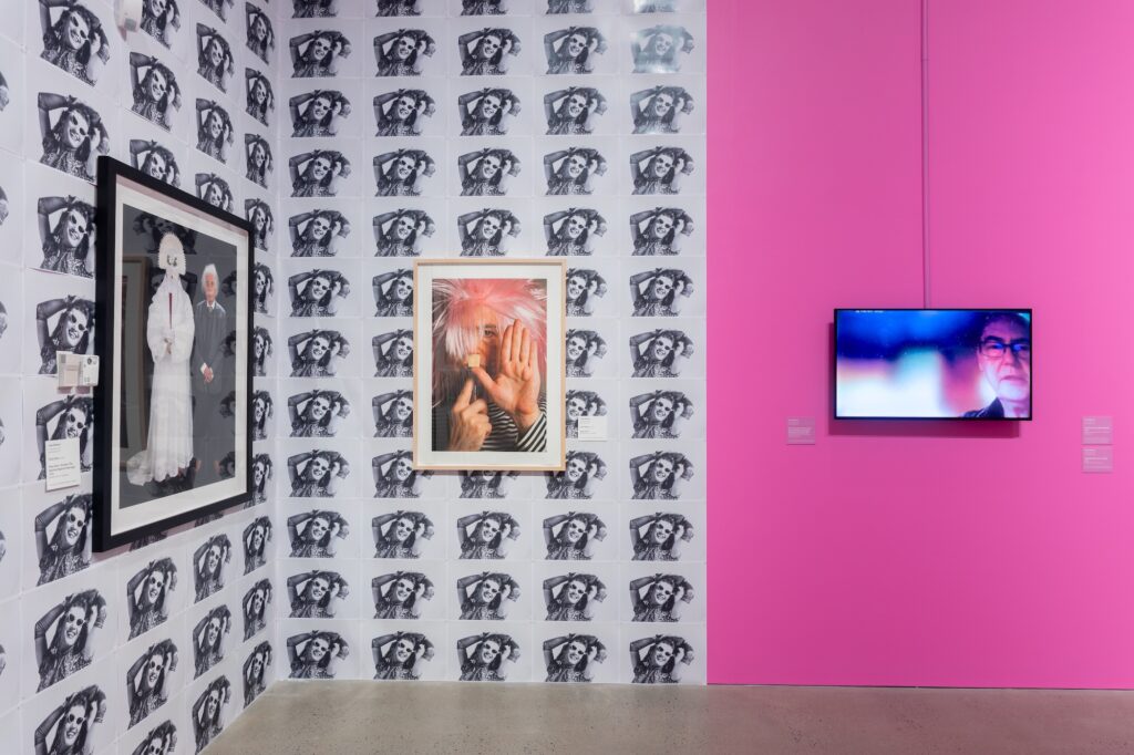 A corner of a gallery shows repeated black and white photographs, a self-portrait of the artist in a pink wig, and a video with the artist against a pink background.