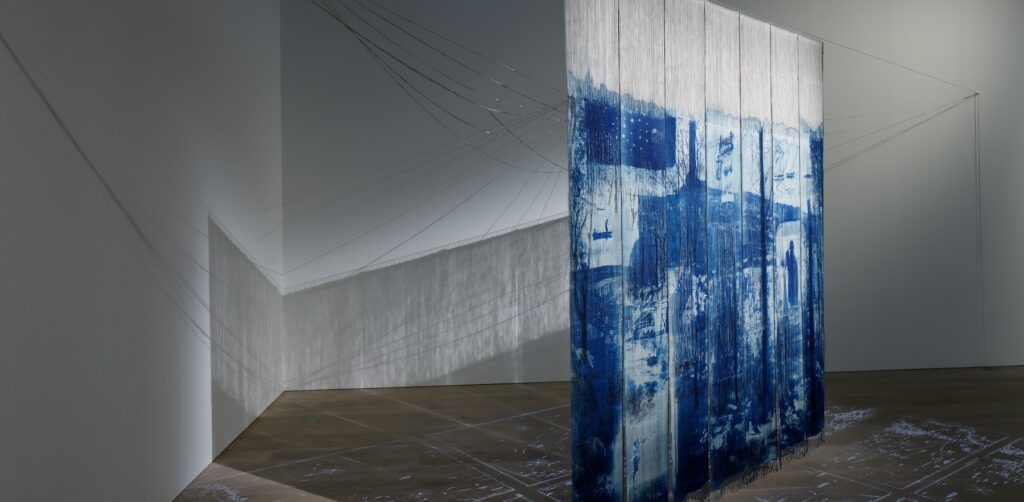 A blue and white textile hangs perpendicular to the corner of a white gallery; shimmery lines and shadows fill the space.