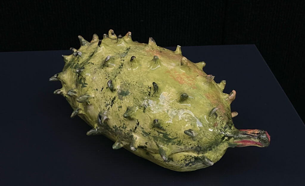 A ceramic of a prickled and green soursop fruit sits on a dark plinth.