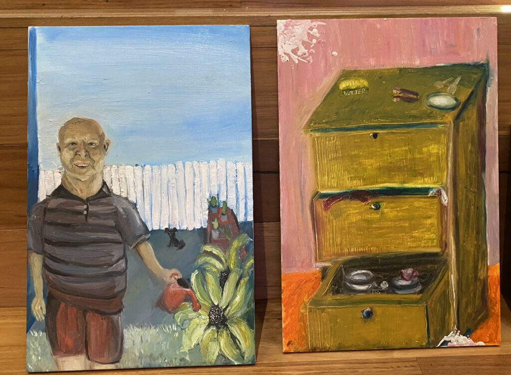 Two small figurative paintings against a wooden background depict an older man watering a garden and a set of bedside drawers.