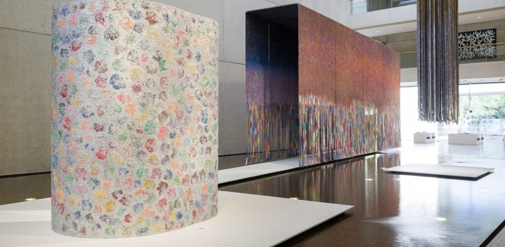 Three large painted installations sit on plinths and hang from above, surrounded by water and inside a concrete gallery. 