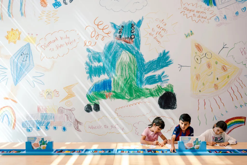 Three children sit at a large table, drawings of monsters and fantastical creatures fill the wall behind them.