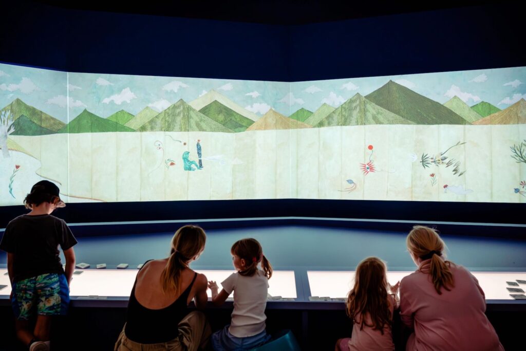 Families sit at a table, a large projected landscape artwork is in front of them.