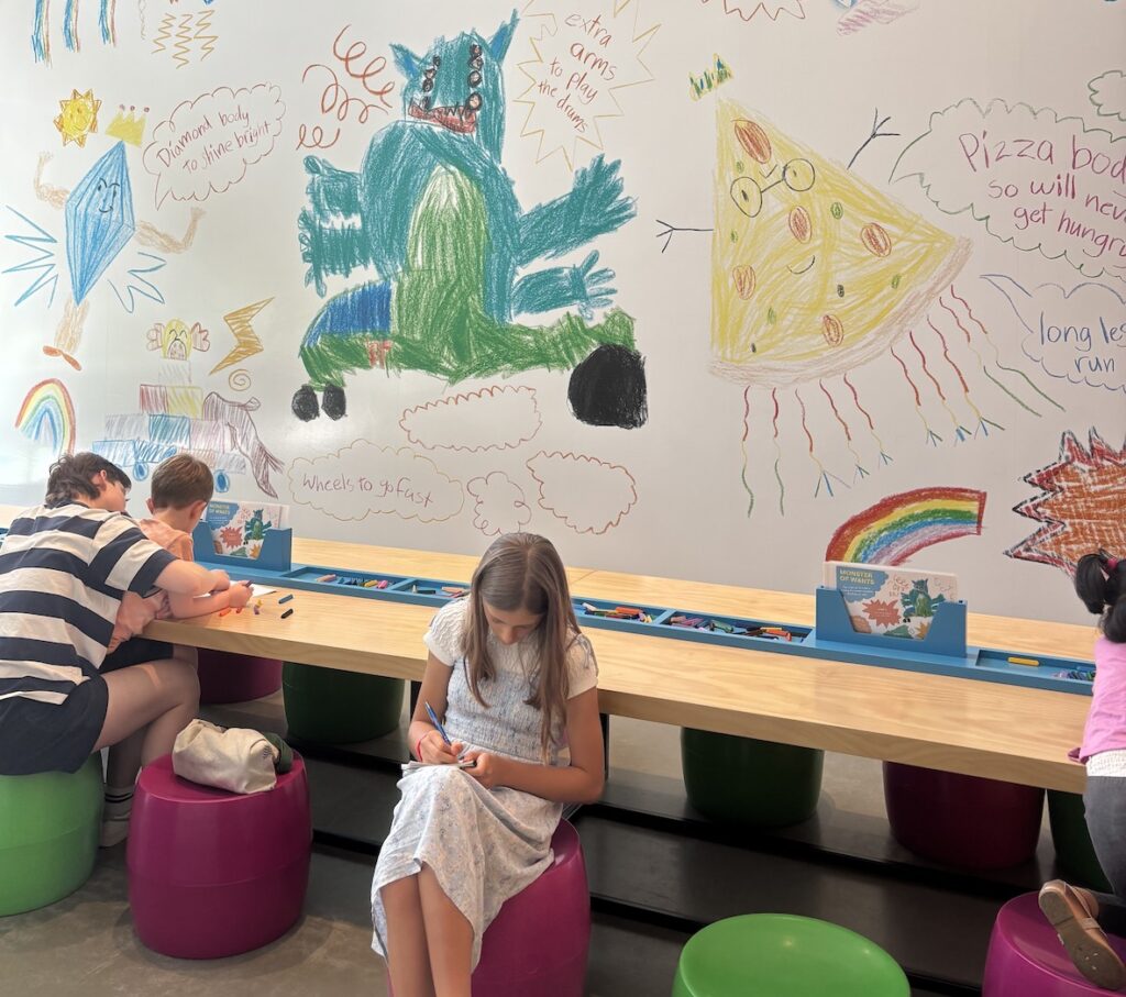 A girl sits front and centre on a stool, head down, writing notes. In the background we can see a white wall covered in childlike drawings of monsters and speech bubbles.