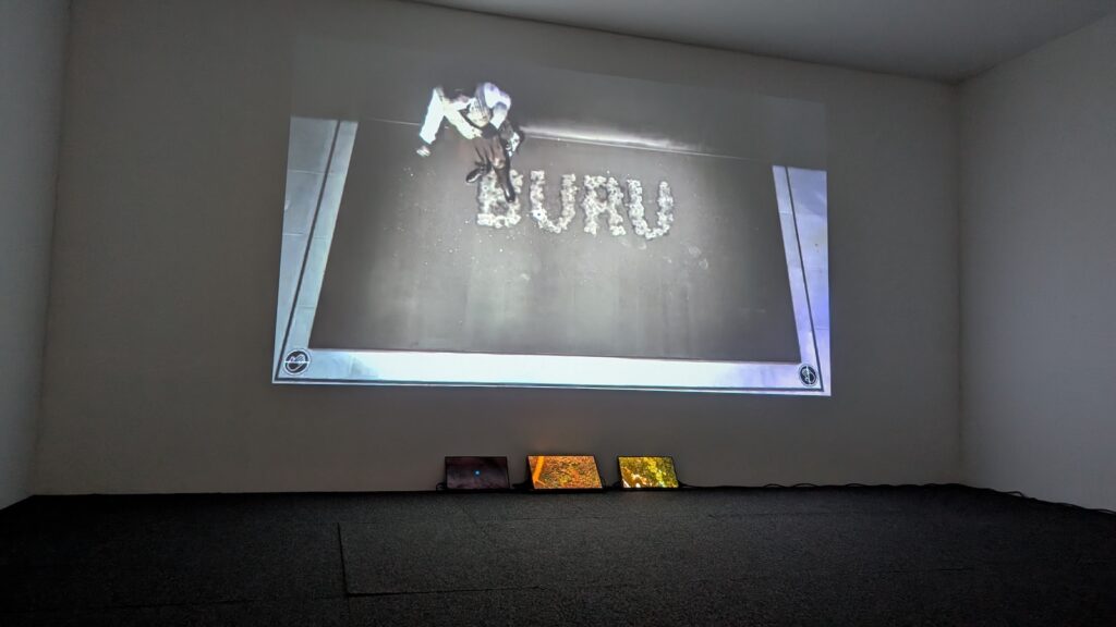 CCTV footage is projected onto a wall. In the footage, a person in a white top walks over an artwork showing the word "buru". Below the projected footage, three screens show overhead drone video of different landscapes.