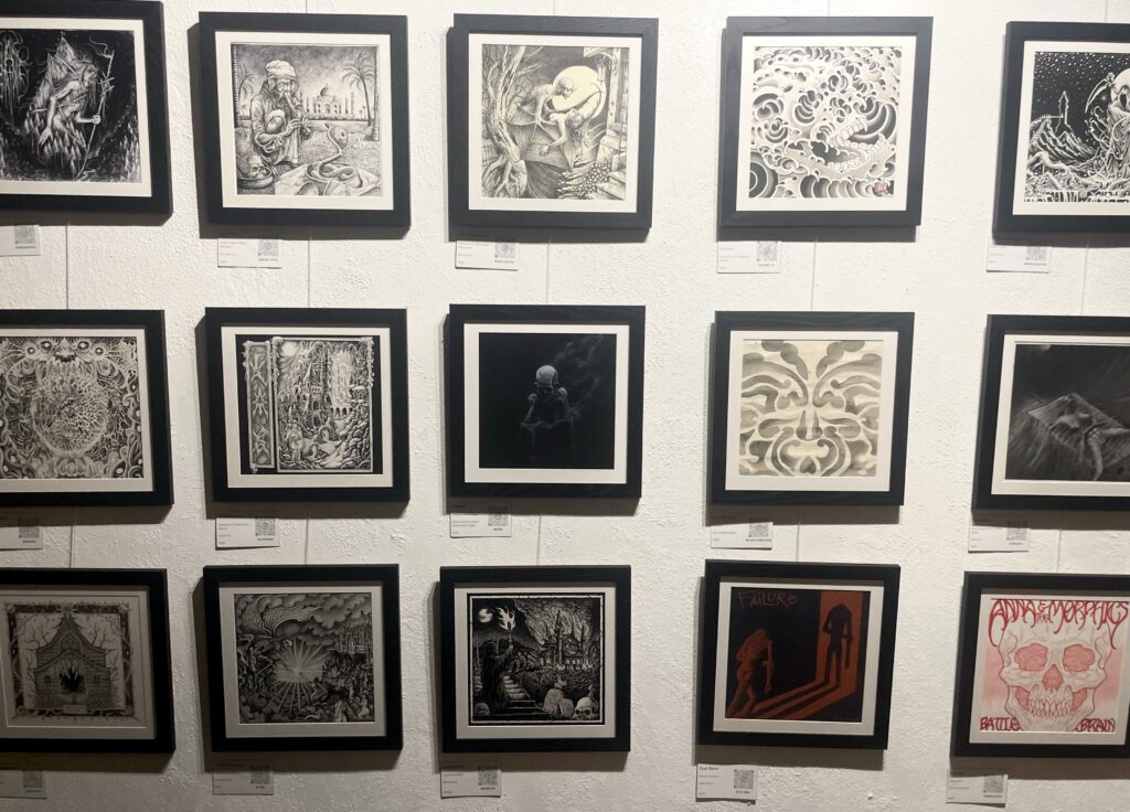 A view of 15 artworks featured in First Pressing, arranged in a grid on a wall. The artworks are all square and in black frames. Most works are greyscale, but one is a stark combination of red and black, while another is in vibrant pink.