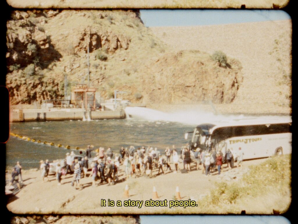 A grainy film still shows a group of people standing in a cluster next to a tour bus and a dam. Text reads: “It is a story about people.”